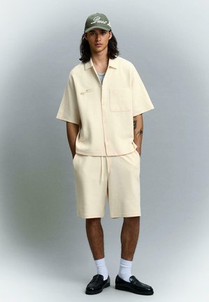 Mladý muž s dlouhými vlasy nosí krémovou krátkou košili na knoflíky a odpovídající šortky, bílé ponožky, černé mokasíny a zelenou baseballovou čepici.