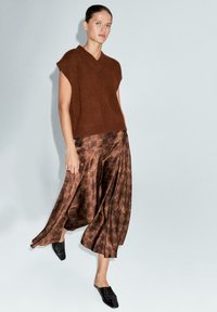 Haut court en tricot marron à col en V, associé à un pantalon large à motifs marron et des chaussures noires à enfiler. Matériaux doux et texturés.
