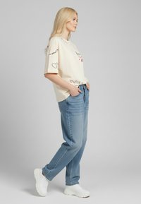 T-shirt crème à manches courtes avec des graphiques ludiques, associé à un jean bleu clair à coupe ample et des baskets blanches, sur un fond neutre.