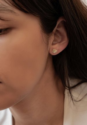 Gold-tone stud earrings shaped like small leaves with a textured surface, set against dark hair and skin, featuring a minimalist design.