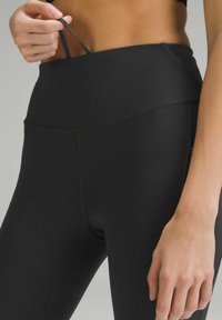 Czarne legginsy z wysokim stanem, wykonane z gładkiego, elastycznego materiału; mają płaski pas i boczną kieszeń dla wygody.