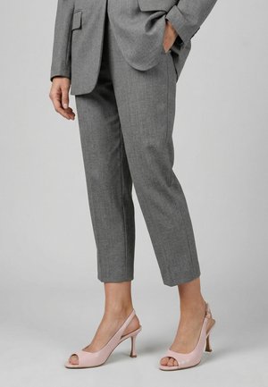 Osoba nosząca krótkie szare spodnie i jasnoróżowe sandały na wysokim obcasie z odkrytymi palcami, stojąca z jedną ręką w kieszeni.