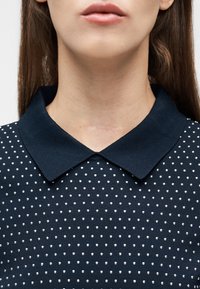 Κάτω μέρος προσώπου και λαιμός γυναίκας με μια αχνή ουλή πάνω από το γιακά ενός μπλε σκούρου πουκάμισου με λευκά πουά.