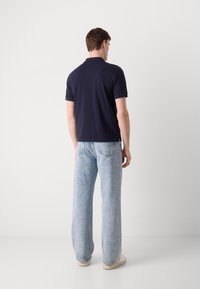 Marinblå pikétröja med korta ärmar som är kombinerad med ljusblå, urtvättade vida jeans. Modellen står vänd bort, iklädd vita skor.
