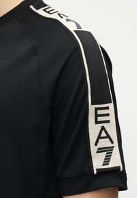 Μαύρο κοντομάνικο πουκάμισο με λευκή και μαύρη ριγέ διακόσμηση στον ώμο που φέρει τα γράμματα "EA7".