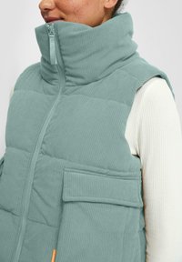 Detail osoby v tyrkysovom ribbovanom prešívanom vestu s vysokým golierom a zipsom, ktorý má na sebe biely dlhý rukáv ribbovaný top.