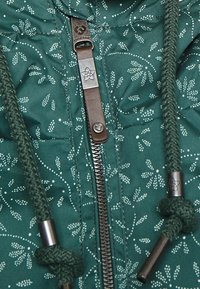 Green quilted jacket featuring floral patterns, a central zipper with a branded pull tab, and drawstring closures with textured ends.
