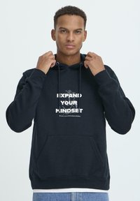 Námornícka mikina s kapucňou vyroben�á z mäkkej látky, s predným vreckom a grafickým textovým dizajnom v bielej farbe, ktorý hovorí "ROZŠÍRTE SVOJE MYSLENIE."