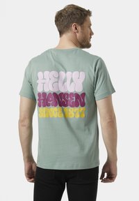 Mintgroene katoenen t-shirt met grote, multikleurige tekst op de achterkant die luidt: "HELly HANSEN SINCE 1877" in speelse lettertypes.