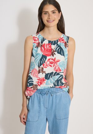 Femme souriante, portant un haut sans manches à fleurs avec des feuilles rouges, bleues et brunes et un pantalon bleu clair à cordon, mains dans les poches.