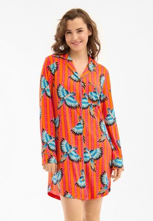 Vrouw glimlachend, draagt een levendige oranje gestreepte overhemdjurk met blauwe en witte vogelprints, staande tegen een effen witte achtergrond.