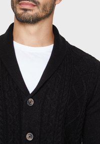 Černý pletený cardigan s texturovaným kabelkovým vzorem, šálovým límcem a třemi knoflíky. Nosený přes bílý tričko, vytvářející vrstvený vzhled.