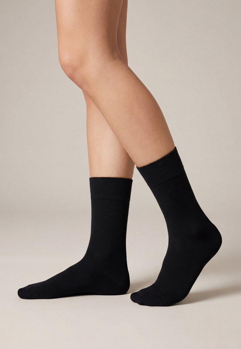 Noge u jednostavnim crnim čarapama do sredine lista na neutralnoj pozadini, jedna stopala ravno na podu, a druga lagano podignuta na prstima.