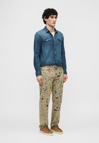 Jeune homme debout, portant une chemise en jean bleue, un pantalon beige à motif floral et des baskets en daim marron, sur un fond clair uni.