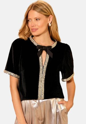 Žena u crnoj baršunastoj kratkoslojnoj majici s ukrasima od šljokica i mašnom, uparena sa sjajnim metalik zlatnim hlačama, pozira s jednom rukom u džepu.