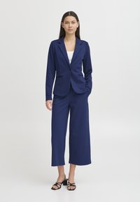 Blazer entallado azul marino con cierre de un solo botón, acompañado de pantalones anchos azul marino. El modelo lleva una blusa blanca y sandalias negras con tiras.