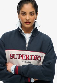 Sudadera navy con cremallera y franjas rojas y crema, con el logo de "SUPERDRY". Lleva una camisa de botones azul claro con puños a rayas debajo.
