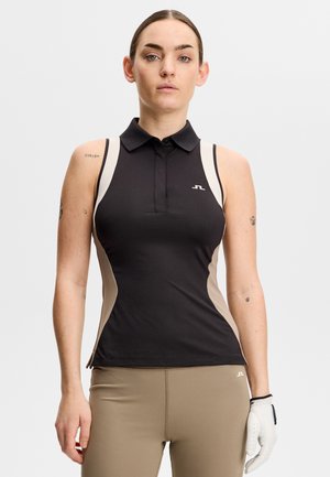 Kobieta ubrana w bezrękawnik polo w kolorze czarno-beżowym i beżowe spodnie, stojąca z białą rękawiczką na prawej dłoni.