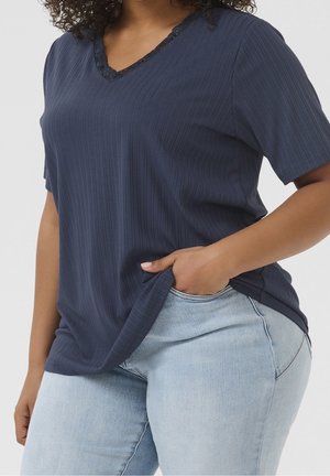 Frau trägt ein marineblaues geripptes T-Shirt mit V-Ausschnitt und Spitzenbesatz sowie hellblaue Jeans, Hand in die Hosentasche gesteckt, vor einem einfarbigen Hintergrund.