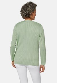 Helles grünes geripptes Strickoberteil mit langen Ärmeln, rundem Halsausschnitt und seitlichen Schlitzen, getragen über weißen Hosen.