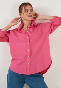 Pink button-up shirt with a relaxed fit, featuring a spread collar and long sleeves, paired with blue denim jeans. Soft cotton fabric.