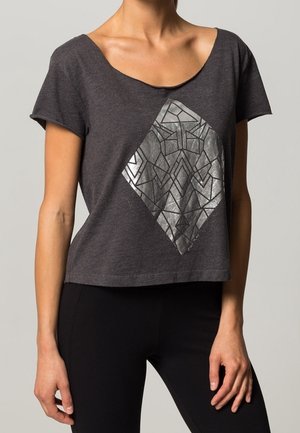 Kobieta nosząca ciemnoszary top z krótkim rękawem z metalicznym, srebrnym geometrycznym diamentowym wzorem, w połączeniu z czarnymi dopasowanymi spodniami.