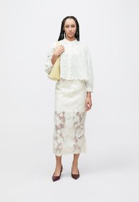 White cropped blouse with eyelet details, layered over a sheer, floral lace skirt. Model holds a soft yellow bag and wears maroon heels.