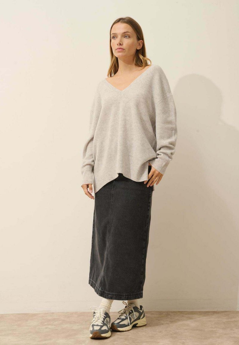 Lichtgrijze oversized trui met een V-hals en ribbeltextuur, gecombineerd met een lange zwarte spijkerrok en meerkleurige sneakers.