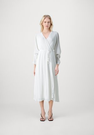 White wrap dress with long, puffed sleeves, textured fabric, a cinched waist, and a flowing hem, accessorized with black strappy heels.