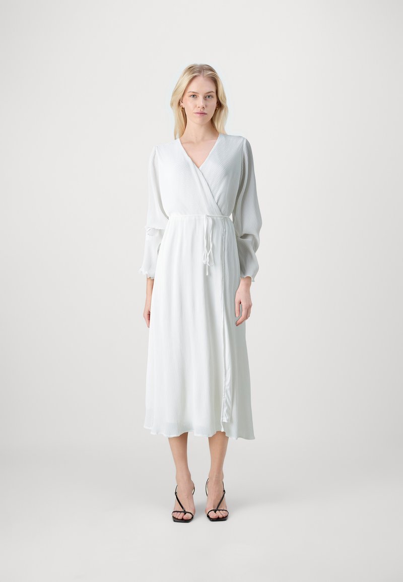 Fehér, hosszú, puffos ujjú, texturált anyagú, deréknál megkötött, és bő hemline-nel rendelkező ruha, fekete pántos magassarkú cipőkkel kiegészítve.