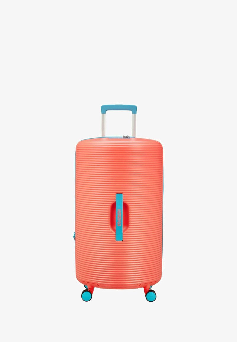 Valise rigide de couleur corail vif avec texture rainurée, poignée accentuée en bleu, poignée rétractable et roulettes pivotantes pour une meilleure mobilité.