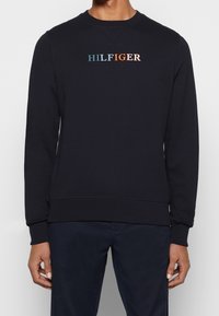 Marinblå sweatshirt med rund halsringning, ribbade muddar och fåll. Har en broderad, flermönstrad "HILFIGER"-logotyp på bröstet. Mjuk textur.