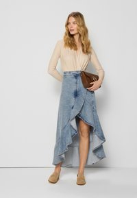 Denim maxi rok met een high-low zoom, gerimpelde rand, lichtblauwe kleur, gecombineerd met een beige wikkelbodysuit en een bruine crossbody tas.