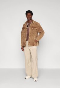 Giacca di velluto a coste marrone con due taschini sul petto, indossata sopra una camicia bordeaux. Pantaloni beige, sneakers bianche. Sfondo semplice.