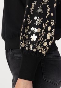 Svart hettegenser med strukturert, elastisk mansjett som har metallisk gullblomsterdesign. Glatt stoff med samlet detalj ved ermet.