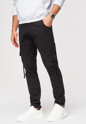 Mężczyzna w czarnych spodniach typu cargo, białych tenisówkach, szarej bluzie, z jedną ręką w kieszeni i srebrnym zegarkiem na lewym nadgarstku.