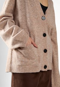 Cardigan beige chiaro con scollo a V, bottoni neri, polsini a costine e tasche laterali. Tessuto morbido, design casual e vestibilità rilassata.