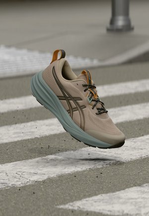 Chaussure de course beige et grise avec des accents orange flottant au-dessus d'un passage piéton sur une route asphaltée.