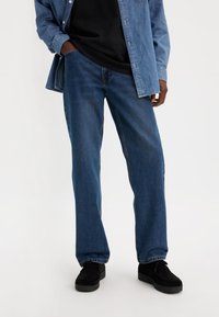 Jean en denim bleu foncé, coupe droite, porté avec une chemise noire et une chemise boutonnée bleu clair. Des chaussures en daim noir complètent la tenue.