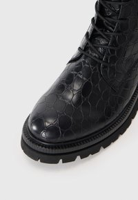 Botte noire à la cheville en cuir texturé avec un motif circulaire, dotée d'une semelle épaisse et robuste et d'un système de laçage noir assorti.