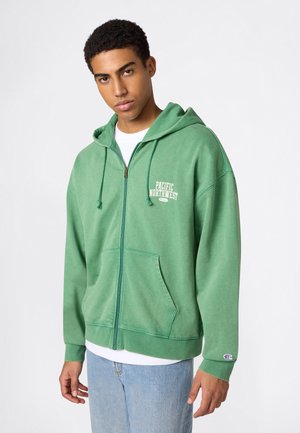 Junger Mann trägt eine grüne Kapuzenjacke mit Reißverschluss und dem Schriftzug "Pacific Northwest", ein weißes T-Shirt und hellblaue Jeans vor einem einfarbigen Hintergrund.