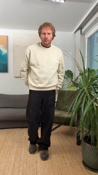 Bézs pulóver kerek nyakkivágással, fekete bordázott nadrág és szürke sportcipő. Beltéri környezet, háttérben növénnyel és absztrakt művészettel.