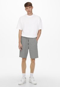 T-shirt bianca a maniche corte e pantaloni corti grigi in maglina con vita elastica, dotati di tasche laterali e cordino, indossati con sneaker bianche.