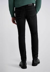 Schwarze Slim-Fit-Jeans mit glatter Textur, ausgestattet mit Gesäßtaschen und einem Etikett am Bund. Kombiniert mit schwarzen, polierten Schuhen.