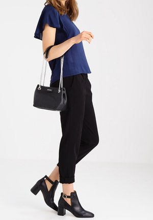 Sac à main noir avec bandoulière en chaîne, blouse navy, pantalon noir court et bottines noires avec design découpé et talon bloc.