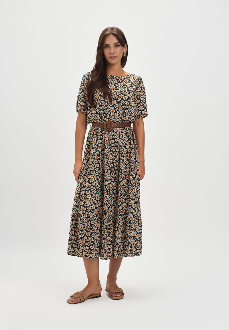 Kwiatowa sukienka midi z ciemnej tkaniny w pomarańczowe i białe wzory, z krótkimi rękawami, z zaakcentowaną talią przy pomocy brązowego paska, zestawiona z beżowymi sandałami.