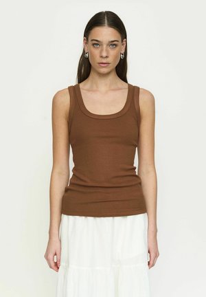 Vrouw met lang bruin haar, zilverkleurige creolen, een bruin mouwloos geribbeld topje en een witte gelaagde rok, staand tegen een effen achtergrond.