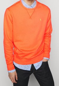 Ljusorange crewneck-tröja med ribbade manschetter och nederkant, lagd över en ljusblå blommig skjorta, stylad med mörka jeans.