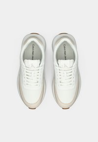 Um par de ténis brancos Calvin Klein Jeans com detalhes em bege, solas grossas e palmilhas com a marca, vistos de cima sobre um fundo branco.