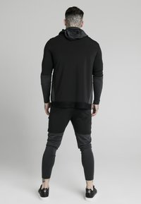 Zwarte sportieve hoodie met een grijze capuchon en accenten, gecombineerd met donkergrijze joggingbroek. Heeft een getailleerd design en ribbelmanchetten.